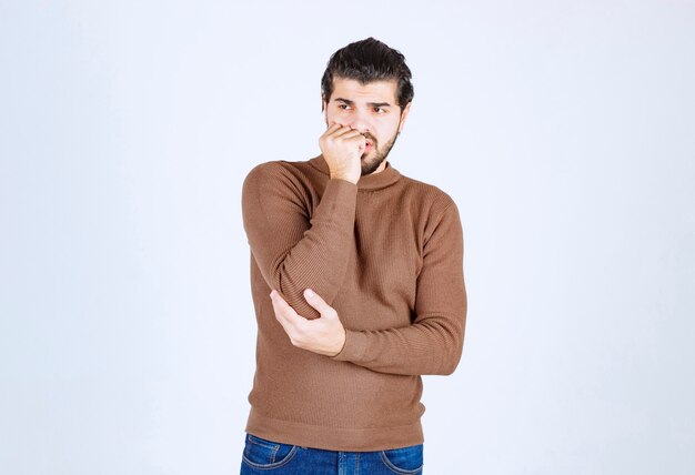 A young man model standing and biting his nails over white wall .