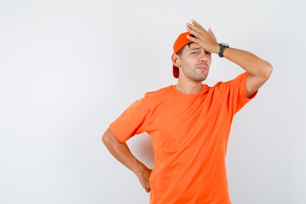 Young man in orange t-shirt and cap holding hand on forehead and looking sorry