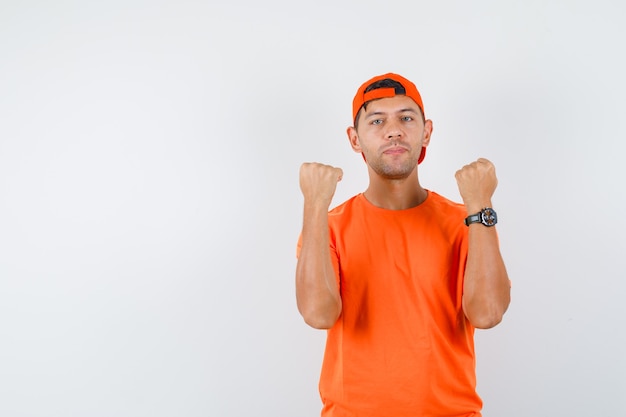 Free photo young man in orange t-shirt and cap holding raised fists clenched and looking confident