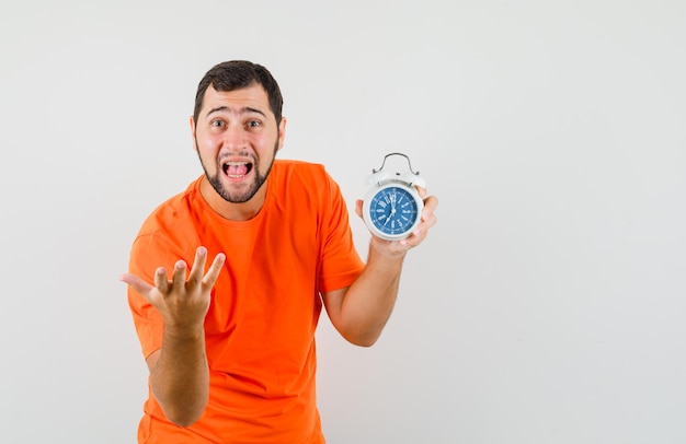 Young man in orange t-shirt holding alarm clock and screaming , front view.