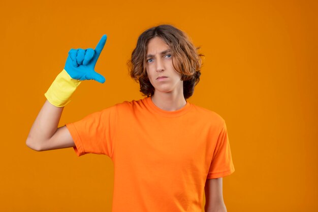 Young man in orange t-shirt wearing rubber gloves gesturing with hand showing small size sign with serious face measure symbol standing over yellow background