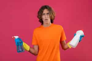 Free photo young man in orange t-shirt wearing rubber gloves holding cleaning spray and bottle of cleaning supplies looking at camera confused spreading hands standing over pink background