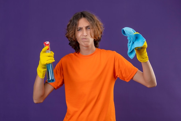 Young man in orange t-shirt wearing rubber gloves holding cleaning spray and rug looking at camera with skeptic expression on face standing over purple background