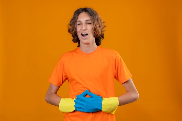 Free Photo young man in orange t-shirt wearing rubber gloves laughing touching his stomach standing over yellow background