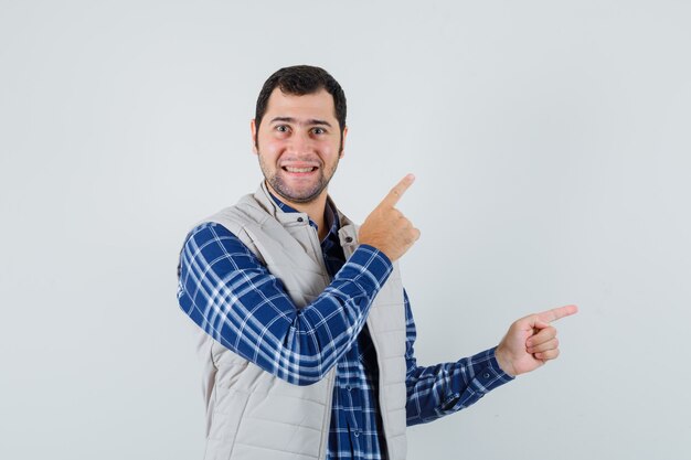 Young man pointing aside in shirt,sleeveless jacket and looking pleased , front view.