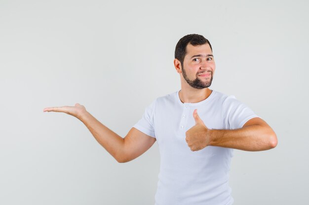 Young man pointing aside while showing thumb up in white t-shirt and looking glad. front view.