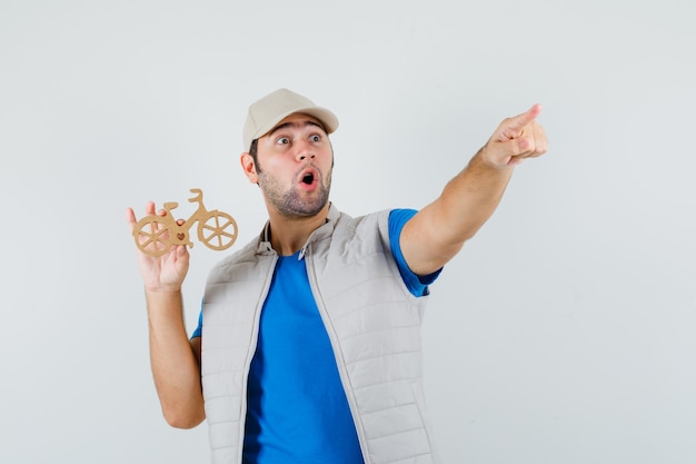 Young man pointing away, holding wooden bike in t-shirt, jacket, cap and looking amazed , front view.