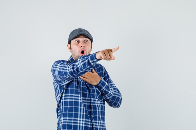 Young man pointing away in shirt, cap and looking surprised. front view.