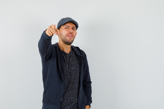 Young man pointing at camera in t-shirt, jacket, cap , front view.