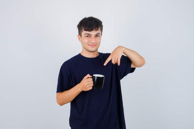 Young man pointing at cup of tea in black t-shirt and looking confident , front view.