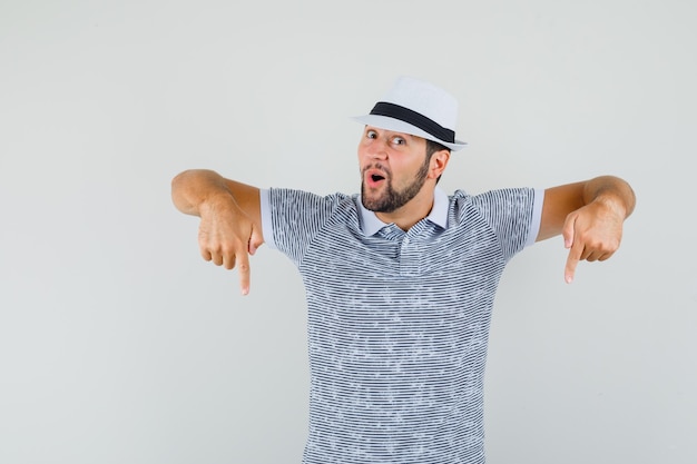 Young man pointing down in striped t-shirt,hat and looking jolly. front view.