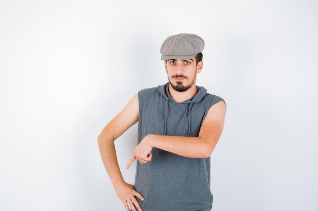 Free photo young man pointing down with index finger in gray t-shirt and cap and looking serious