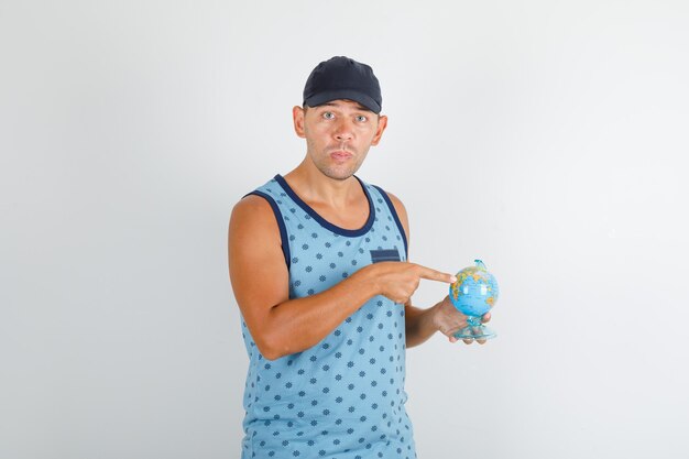 Young man pointing finger at globe in blue singlet with cap and looking confused