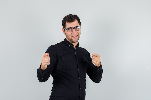 Young man pointing fingers at camera in black shirt, glasses and looking cheerful