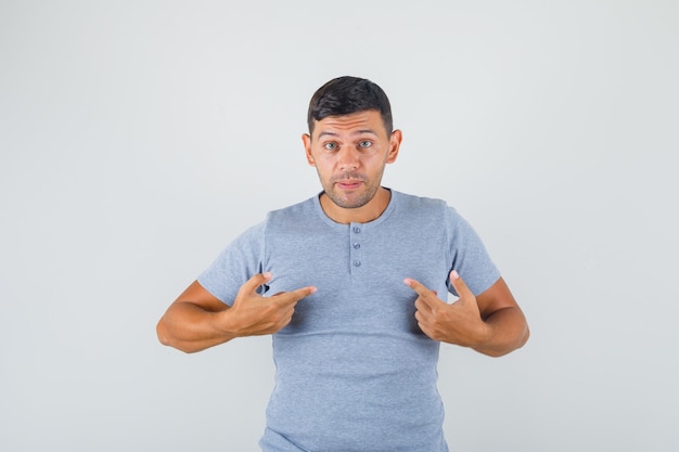 Young man pointing fingers at himself and asking 'me?' in grey t-shirt and looking doubted