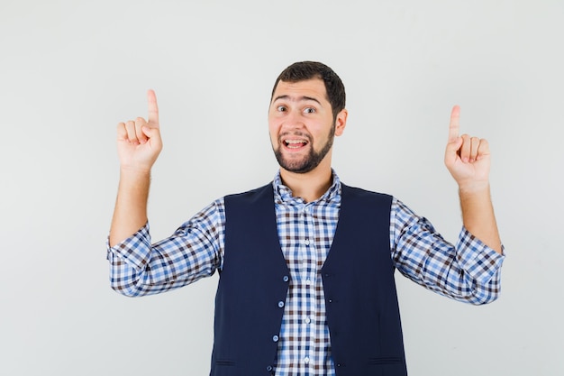 Young man pointing fingers up in shirt, vest and looking cheery , front view.