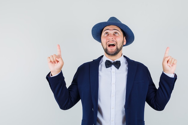 Young man pointing fingers up in suit, hat and looking hopeful , front view.