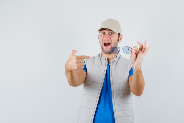 Young man pointing at glasses in t-shirt, jacket, cap and looking glad. front view.