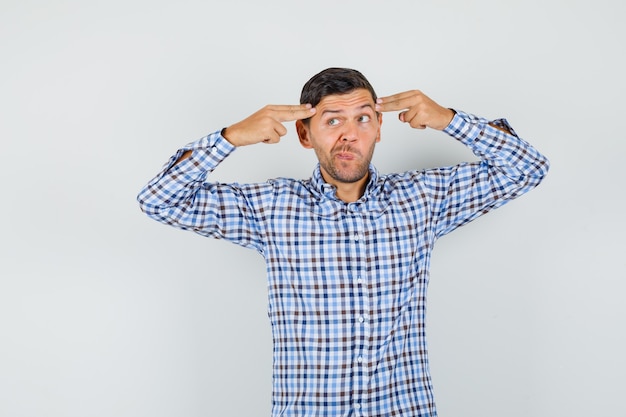 Young man pointing hands and fingers to head in checked shirt and looking pensive.