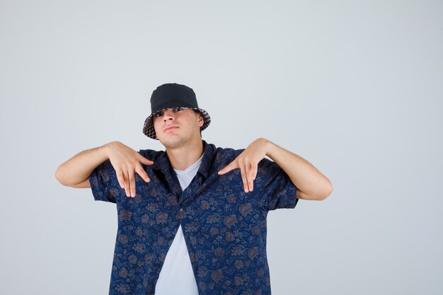 Young man pointing at himself with hands in white t-shirt, floral shirt, cap and looking confident , front view.