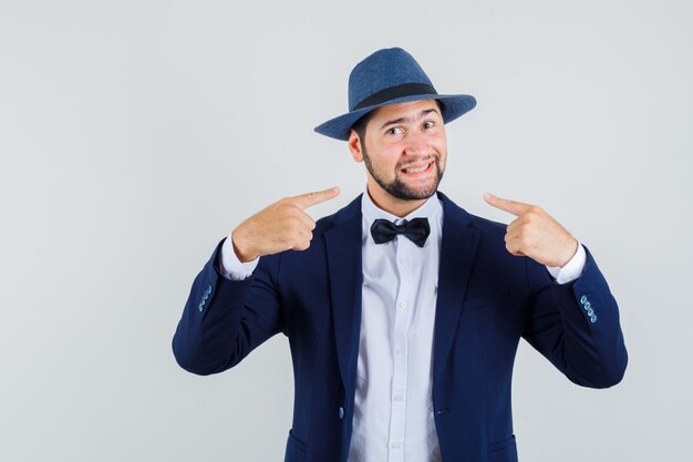 Young man pointing at his smile in suit, hat and looking merry , front view.