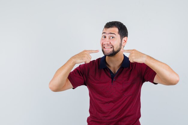 Young man pointing on his teeth in red t-shirt and looking concentrated , front view.