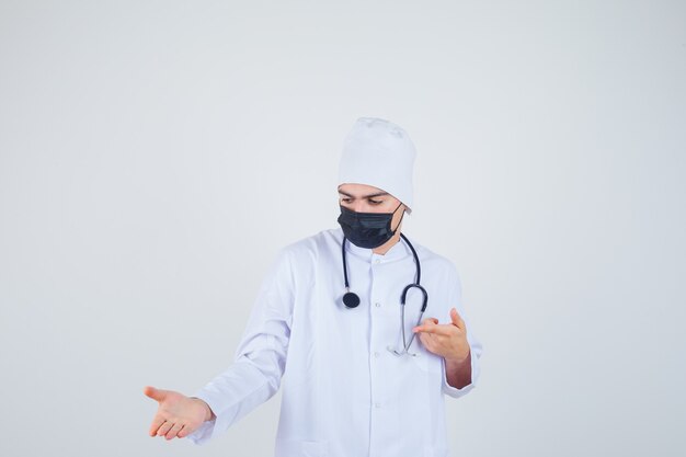 Young man pointing to the left side, pretending to show something in white uniform, mask and looking confident 