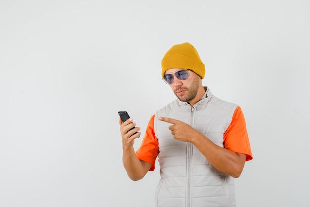 Young man pointing at mobile phone in t-shirt, jacket, hat front view.