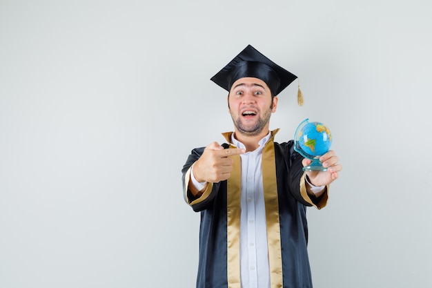 Free Photo young man pointing at school globe in graduate uniform and looking joyful. front view.