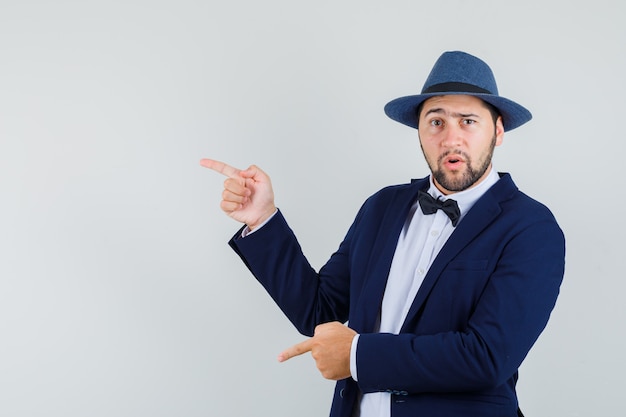 Free photo young man pointing to the side and down in suit, hat and looking confused , front view.