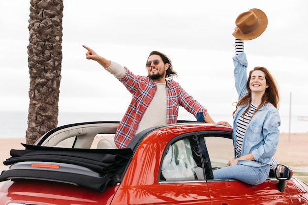 Young man pointing to side near woman waving hand with hat and leaning out from car