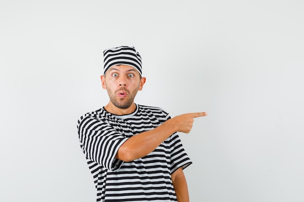 Young man pointing to the side in striped t-shirt hat and looking scared  