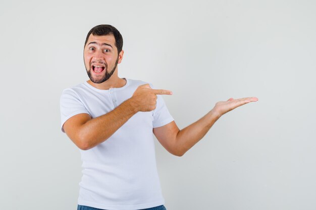 Young man pointing to side in white t-shirt and looking optimistic , front view.