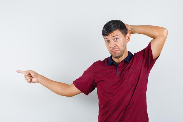 Young man pointing to side with hand behind head in t-shirt and looking hesitant. front view.