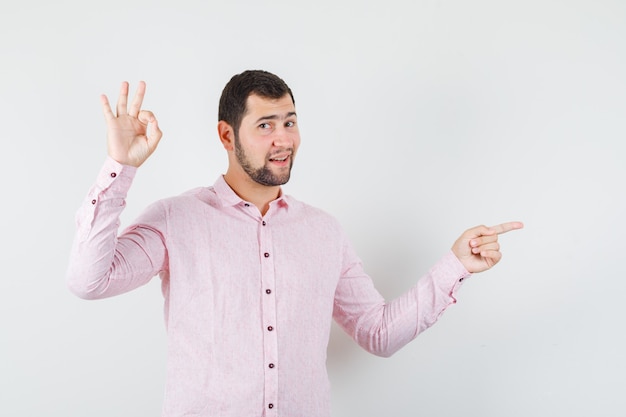 Young man pointing to side with ok sign in pink shirt and looking confident