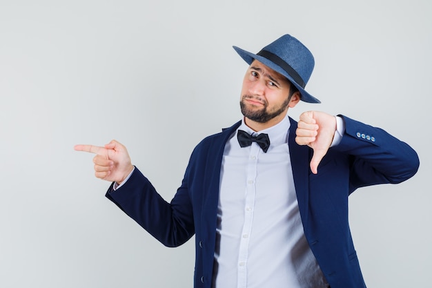 Young man pointing to the side with thumb down in suit, hat and looking displeased. front view.