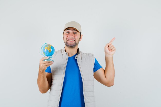 Young man pointing up, holding school globe in t-shirt, jacket, cap and looking glad , front view.