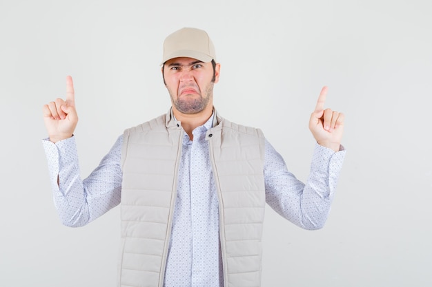 Free photo young man pointing up in shirt,sleeveless jacket,cap and looking dissatisfied. front view.