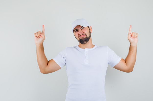 Free photo young man pointing up in white t-shirt,cap and looking disgusted , front view.