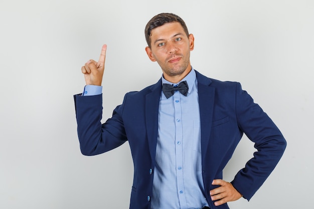 Young man pointing up with hand on waist in suit and looking cheerful 