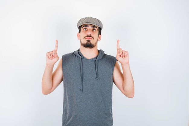 Young man pointing up with index fingers in gray t-shirt and cap and looking serious