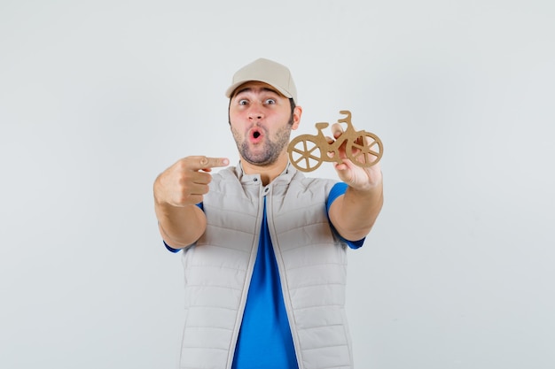 Free photo young man pointing at wooden toy bike in t-shirt, jacket, cap and looking amazed. front view.