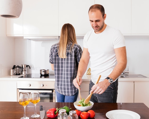 Free photo young man preparing the salad and her wife standing behind him cooking food in the kitchen