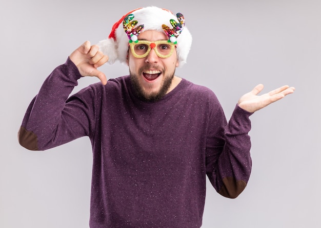 Free photo young man in purple sweater and santa hat wearing funny glasses  happy and joyful smiling cheerfully showing thumbs down standing over white wall