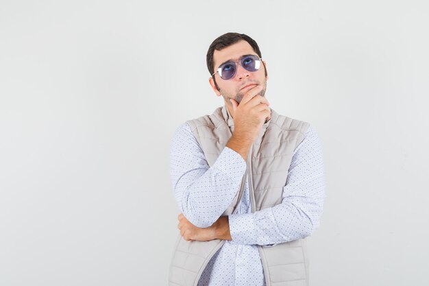 Young man putting on glasses while standing in thinking pose in beige jacket and cap and looking pensive. front view.