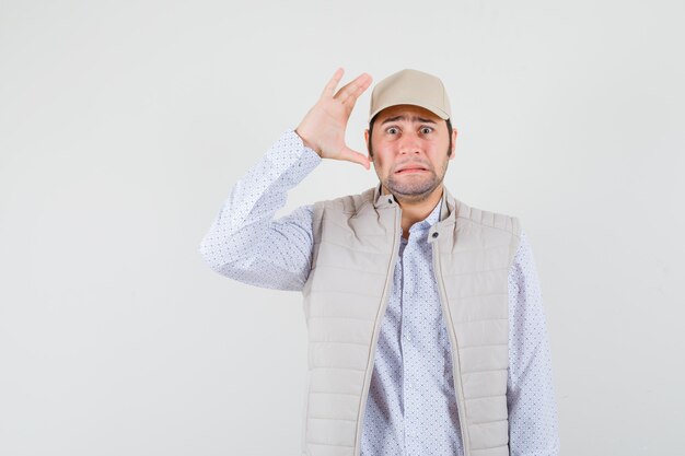 Young man raising hand near ear in beige jacket and cap and looking surprised. front view.