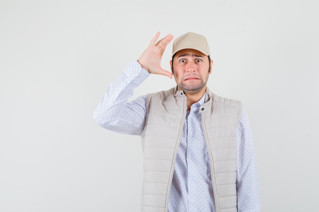 Young man raising hand near ear in beige jacket and cap and looking surprised. front view.