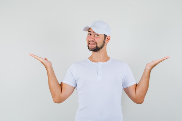 Free Photo young man raising hands like holding or showing something in white t-shirt,cap and looking joyful , front view.