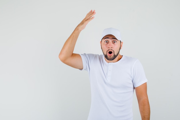 Young man raising his arm with open palm in white t-shirt and looking astonished. front view.