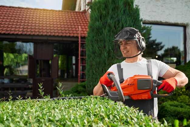 Young man in safety mask and gloves shaping bushes